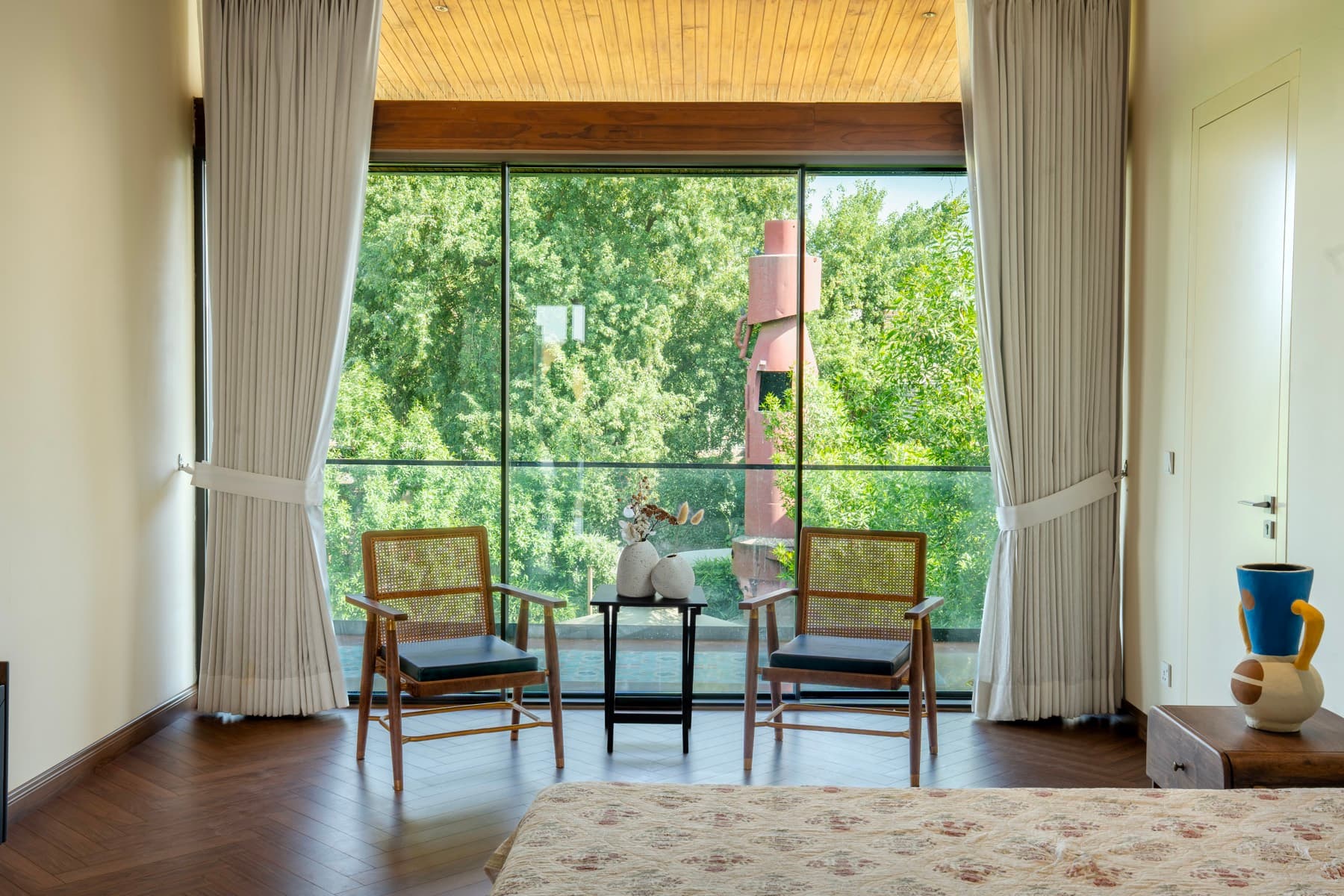 First-floor bedroom with cane armchairs on herringbone timber, the red kupla chimney framed by full-height glass