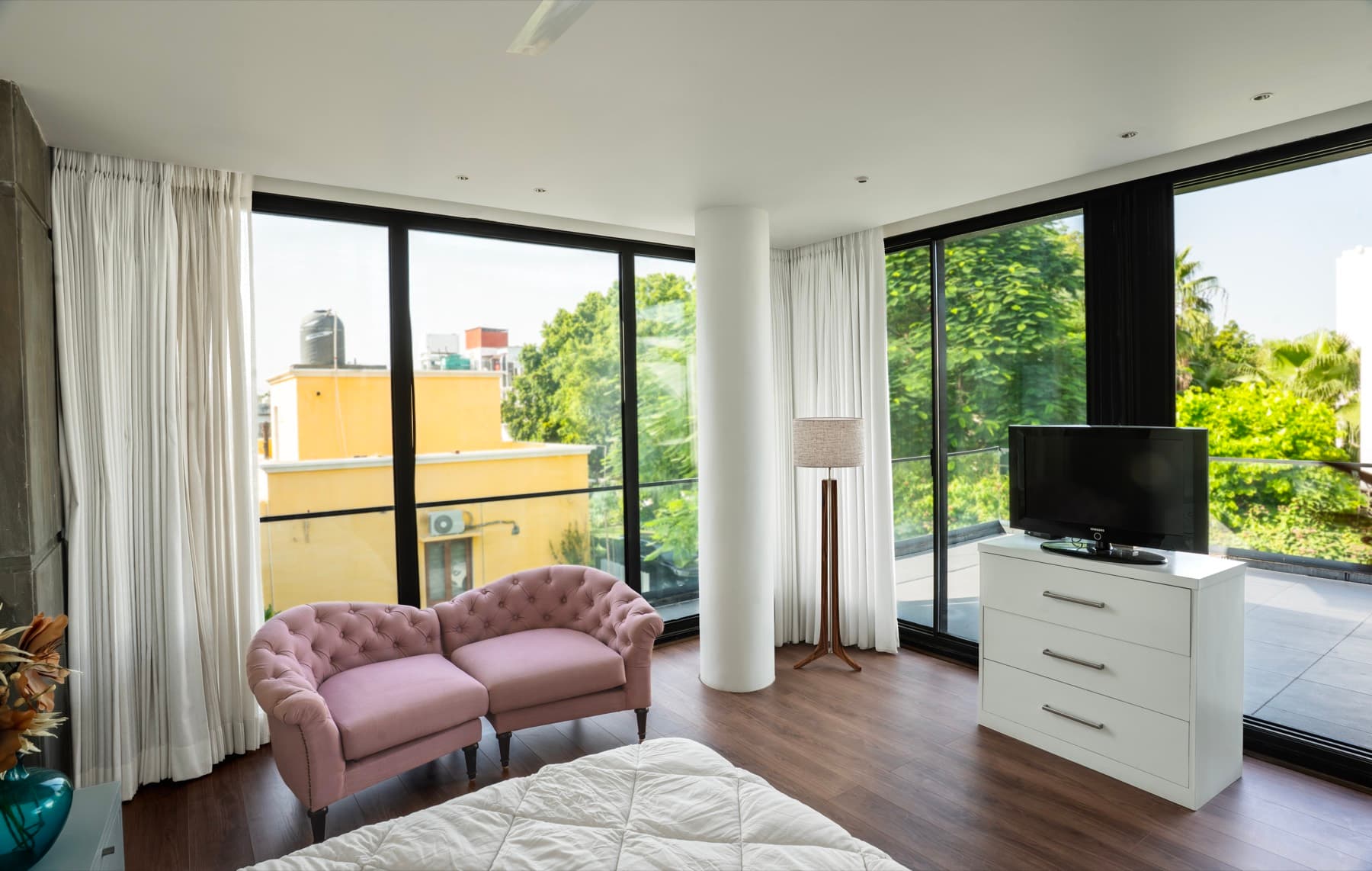 Corner bedroom glazed on two sides around a white column, with pink tufted chairs and terrace beyond