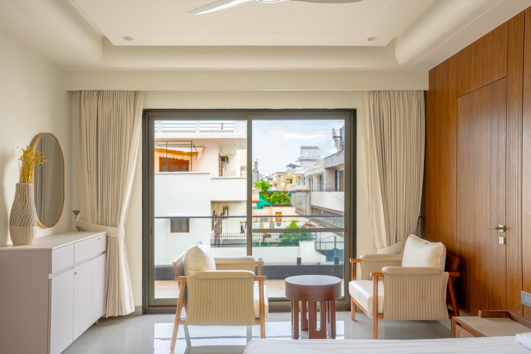Bedroom with two cream armchairs before a sliding door to the balcony, fluted walnut wall alongside