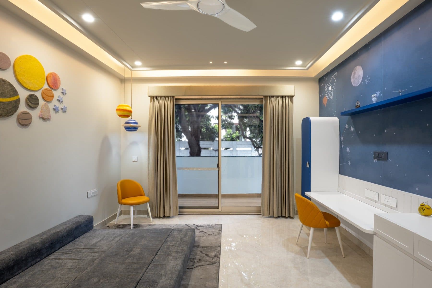 Child's bedroom with a navy starry wall, white study desk, mustard chairs and hanging planet models