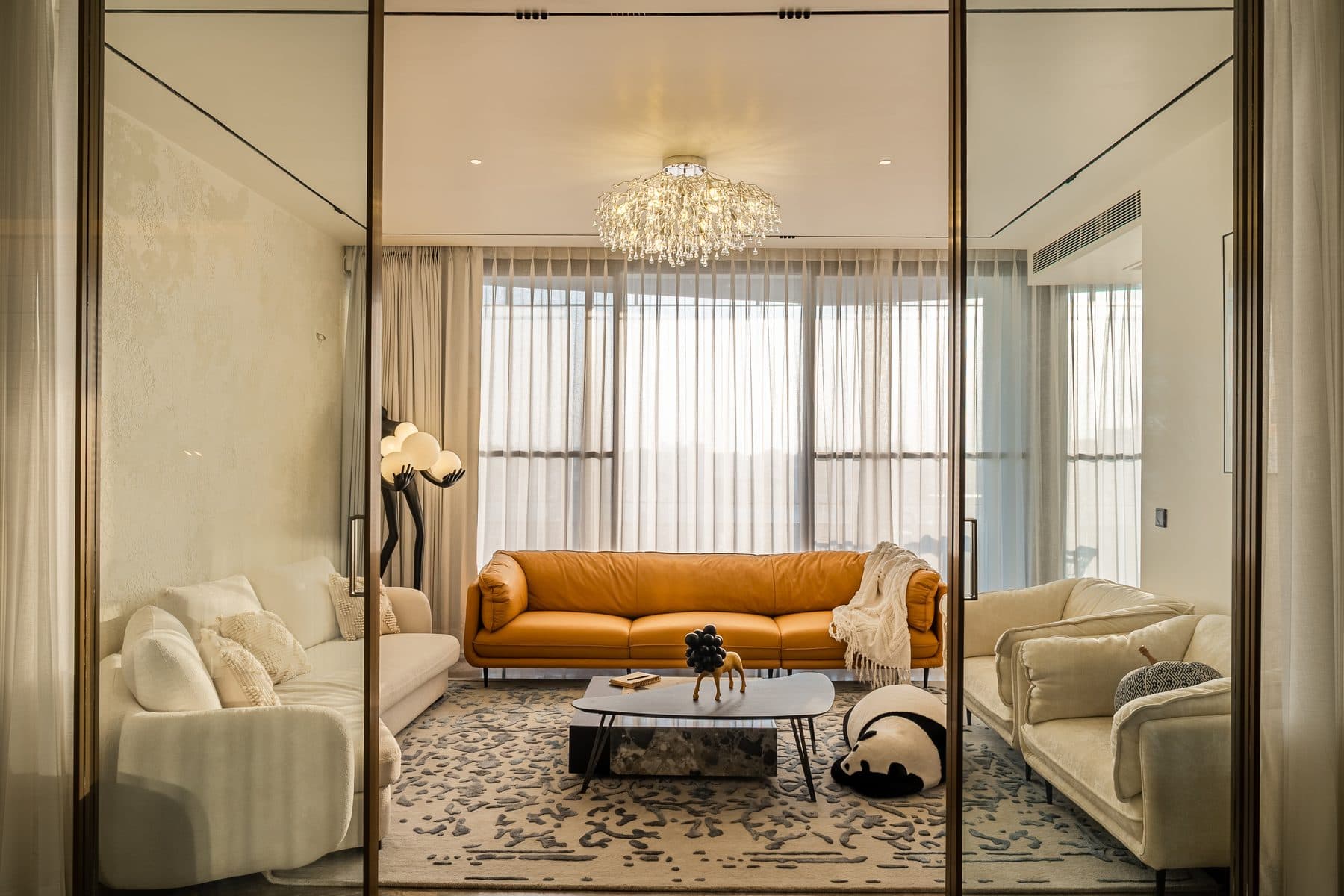 Drawing room seen through gold-framed glass doors: burnt-orange leather sofa, cream boucle seating, crystal chandelier