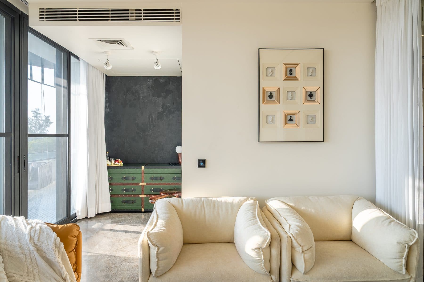 Sitting area with cream tub sofas beneath a framed art grid, the green trunk-style bar chest beyond